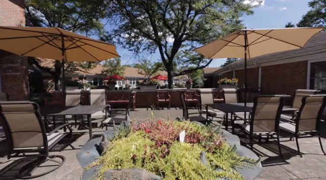 Outdoor patio area with several tables and chairs under large beige umbrellas, surrounded by brick buildings and greenery, with a large tree in the background and a planter with various plants in the foreground.