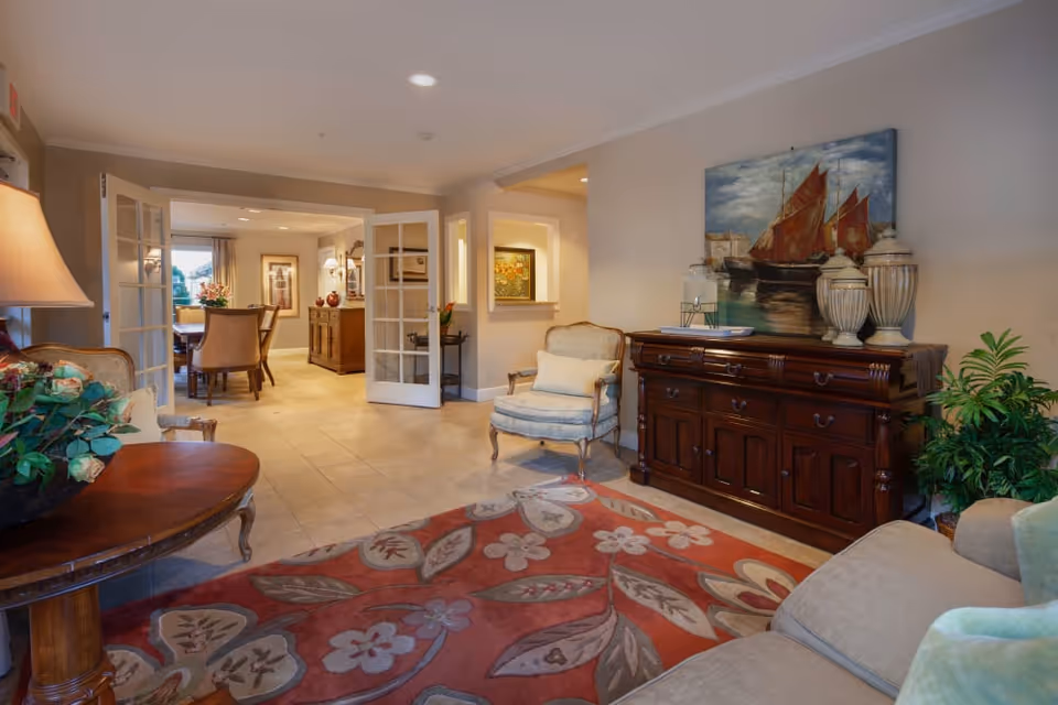 A cozy and well-lit living room area in Cedar Crest Alzheimer's Special Care Center featuring a floral patterned rug, a wooden side table with a flower arrangement, an upholstered armchair with a cushion, a large wooden cabinet with decorative vases and a painting of sailboats above it, and a view into a dining area through open French doors.