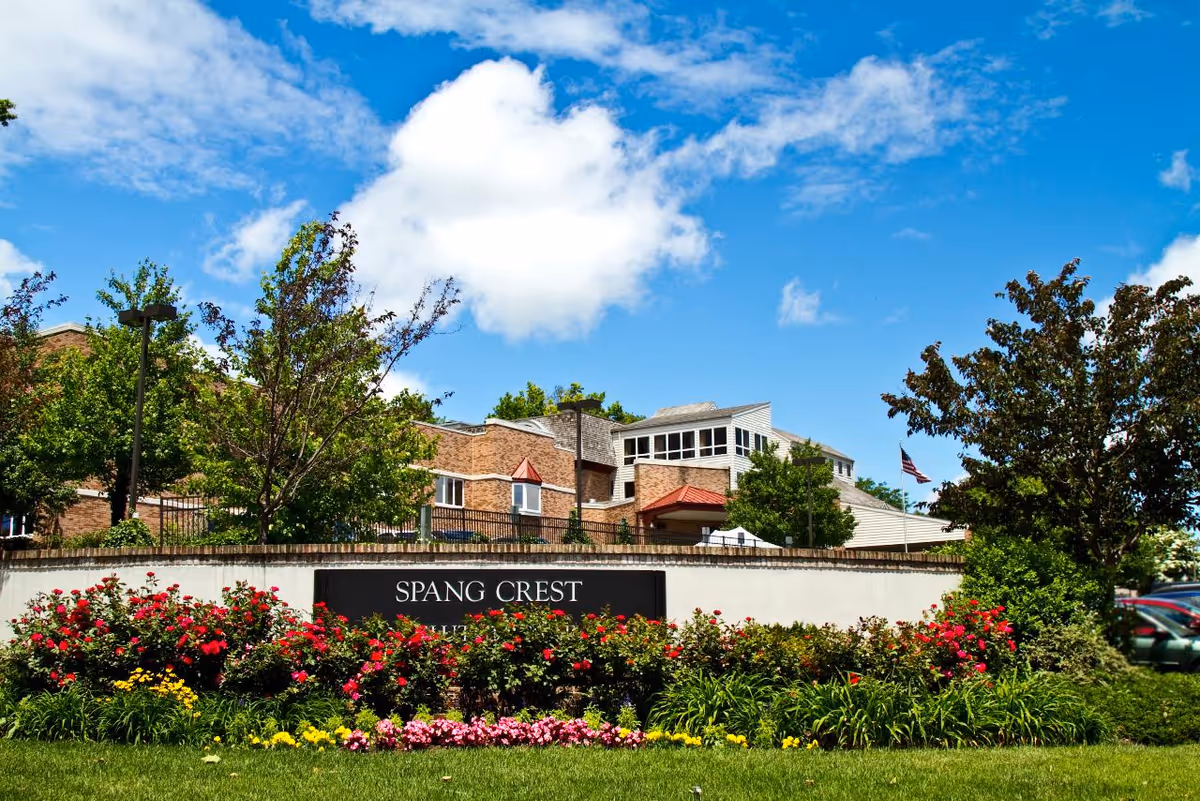 Front view of a brick senior living facility behind a landscaped flowerbed and a sign reading "SPANG CREST" under a blue sky.