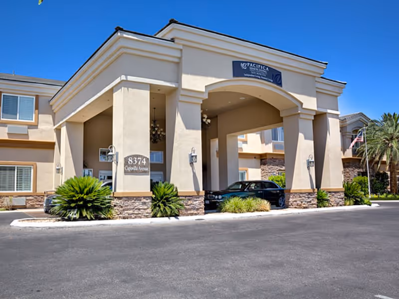 Front entrance and covered porte-cochere of a beige senior living facility with an address plaque reading 8374 and a parked car.