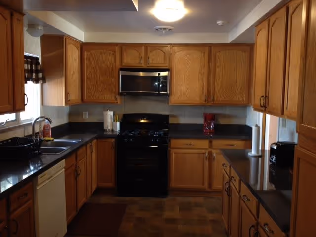 A kitchen with wooden cabinets, black countertops, a black stove with a microwave above it, a dishwasher, a sink under a window with a checkered valance, and various kitchen items such as paper towels, a toaster, and a coffee maker.