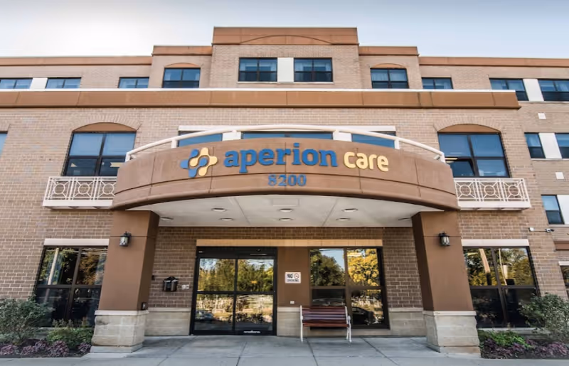 Front exterior view of a multi-story brick building with a curved entrance canopy displaying the sign 'aperion care 8200'. There are large windows on each floor and a bench near the entrance door.