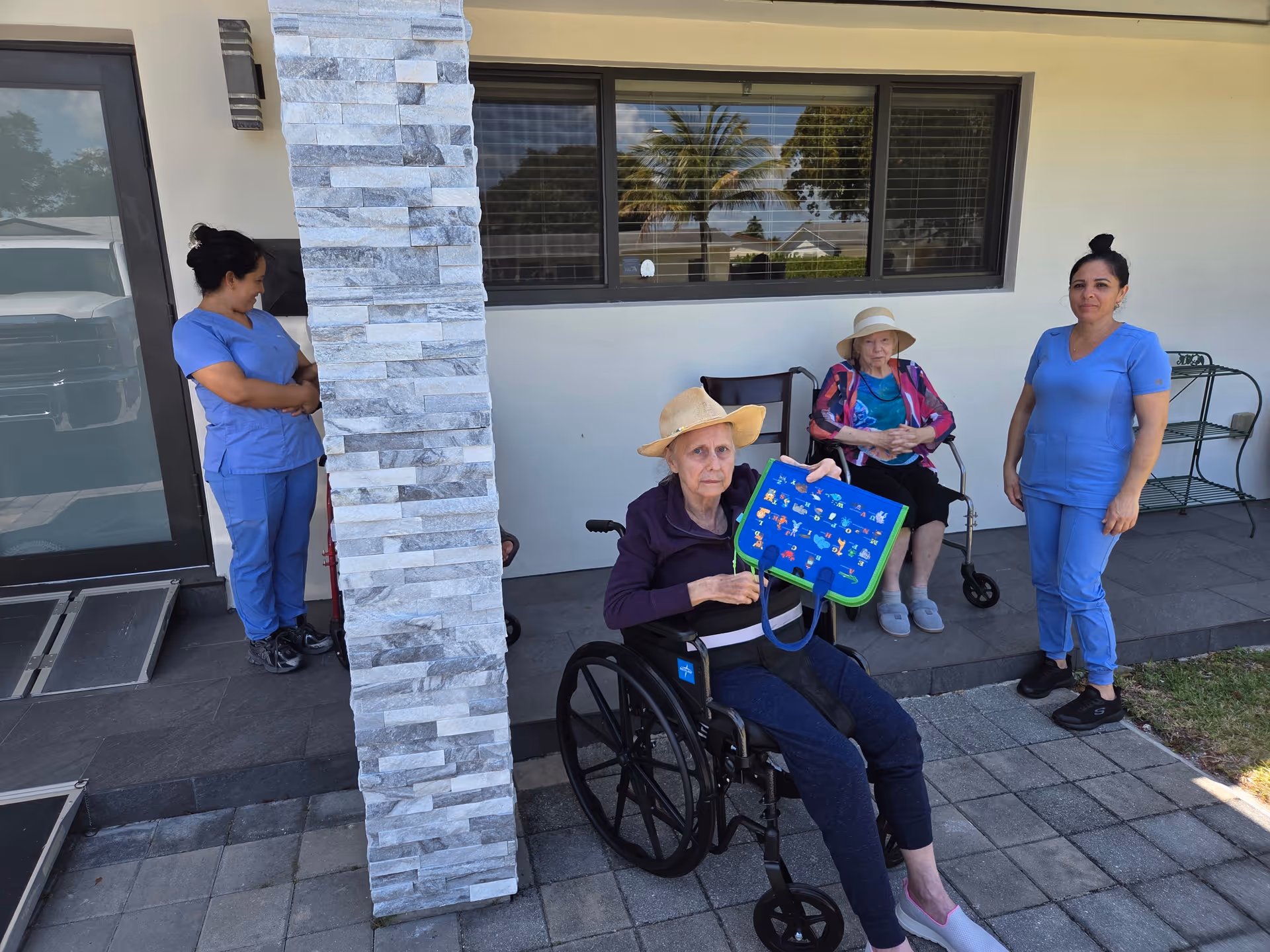 Two elderly women sitting outside a building, one in a wheelchair holding a colorful bag, and the other seated on a chair. Two caregivers in blue scrubs stand nearby, one leaning against a stone pillar and the other standing on the right. The setting appears to be a patio area with a window and a door in the background.