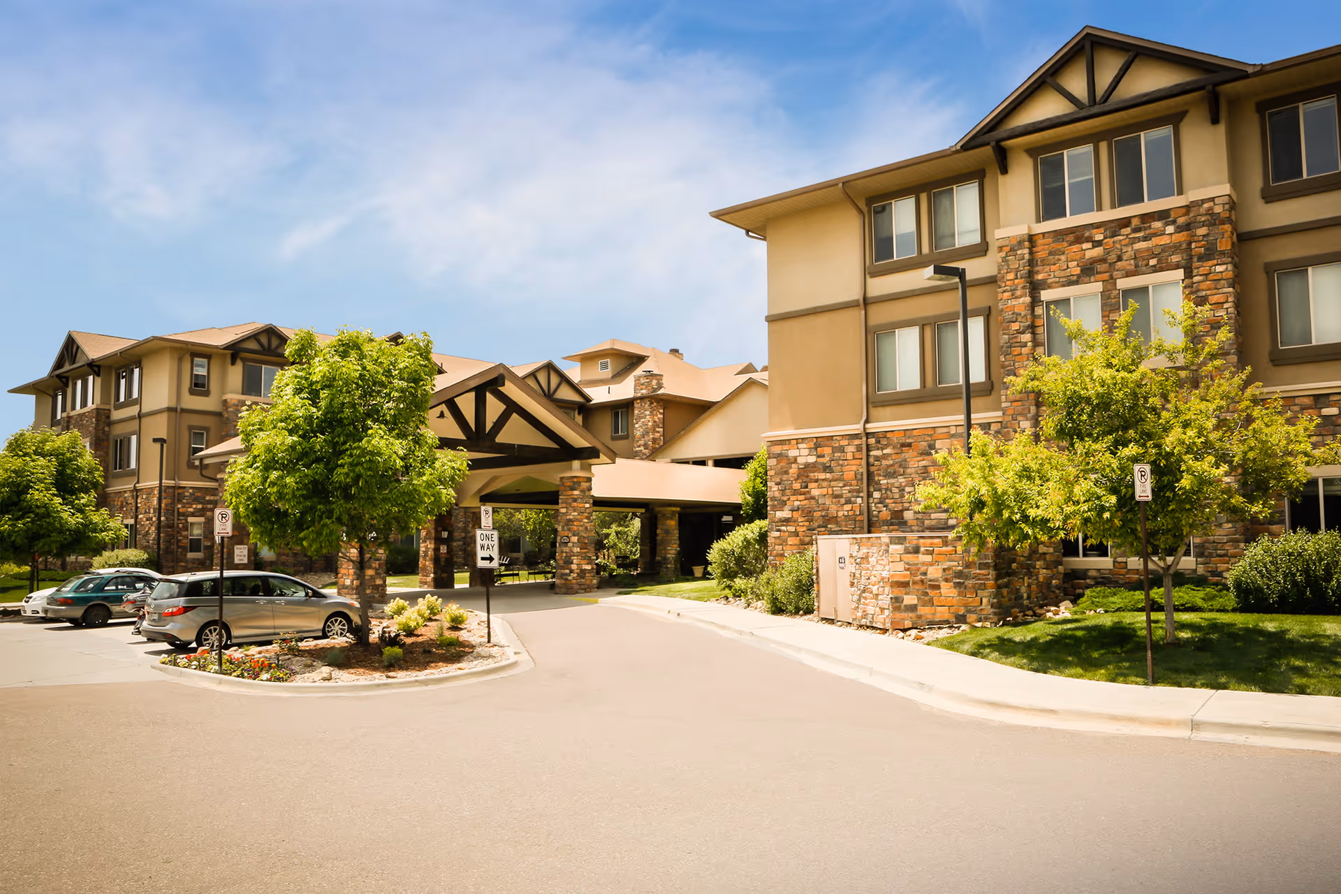 Exterior view of a senior living facility named Bethesda Gardens showing a multi-story building with stone and beige siding, a covered entrance with wooden beams, several trees, parked cars, and a clear blue sky.