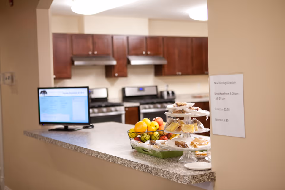 Interior view of a kitchen area with dark wooden cabinets, two stainless steel stoves with range hoods, and a countertop in the foreground displaying a tiered tray of assorted pastries covered in plastic wrap and a basket of fresh fruit. A small monitor screen is also on the counter, and a printed dining schedule is posted on the wall.