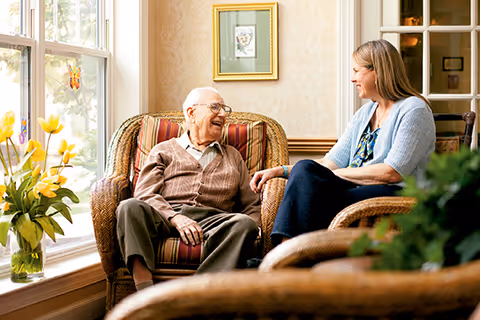 An elderly man and a woman sitting in wicker chairs in a cozy, well-lit living room with large windows and floral decorations, engaged in a cheerful conversation.