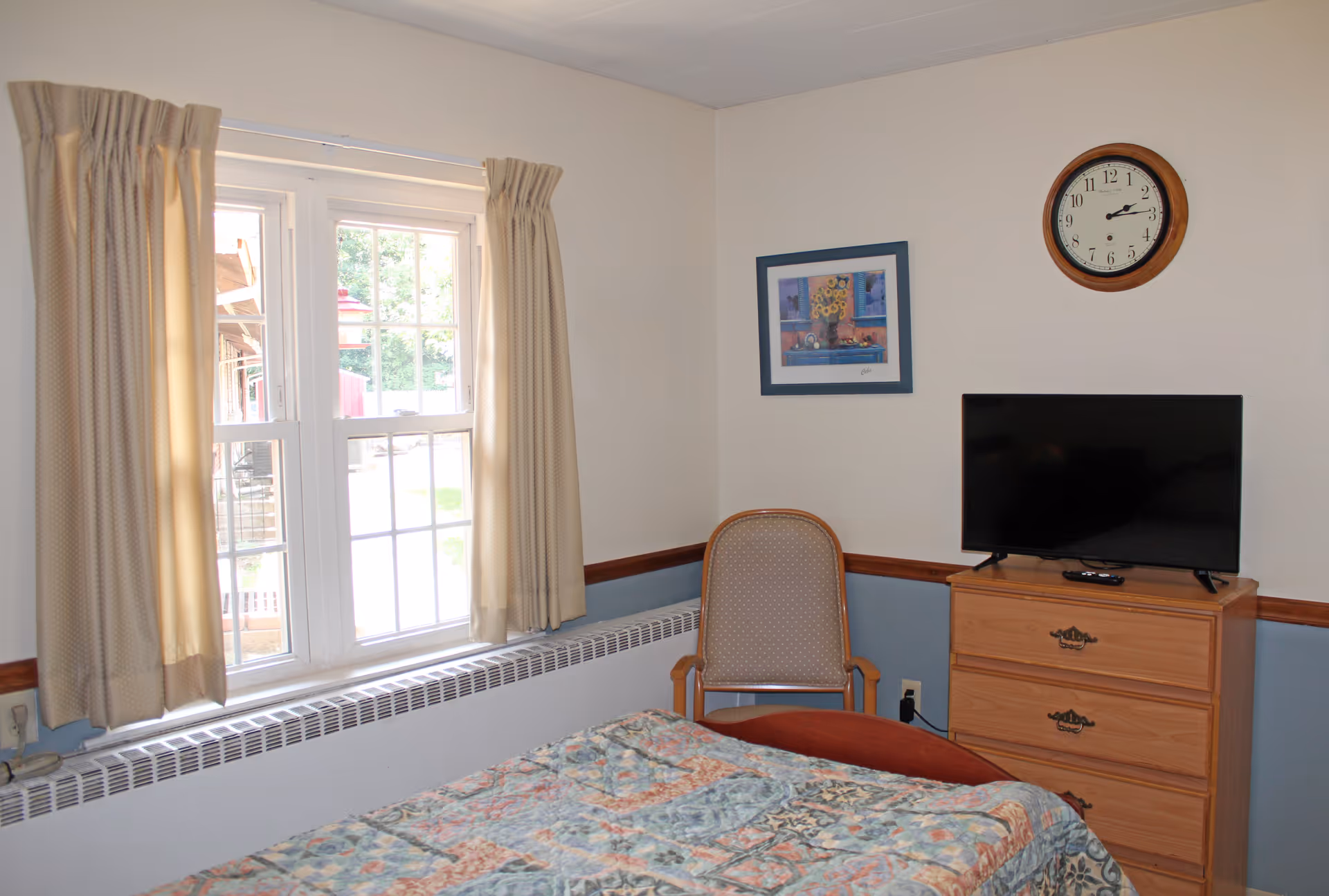 A small bedroom with a bed covered in a patterned quilt, a wooden dresser with a flat-screen TV on top, a cushioned chair, a wall clock, a framed picture, and a window with beige curtains letting in natural light.