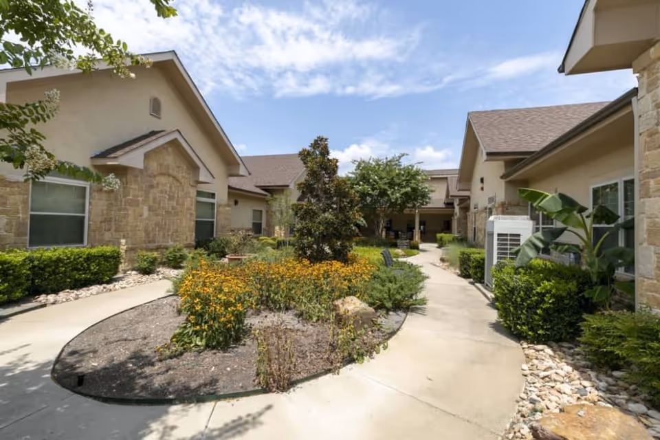 Outdoor courtyard area of a senior living facility with a paved walkway, landscaped garden beds with yellow flowers, shrubs, and trees, surrounded by single-story buildings with stone and stucco exteriors under a partly cloudy sky.