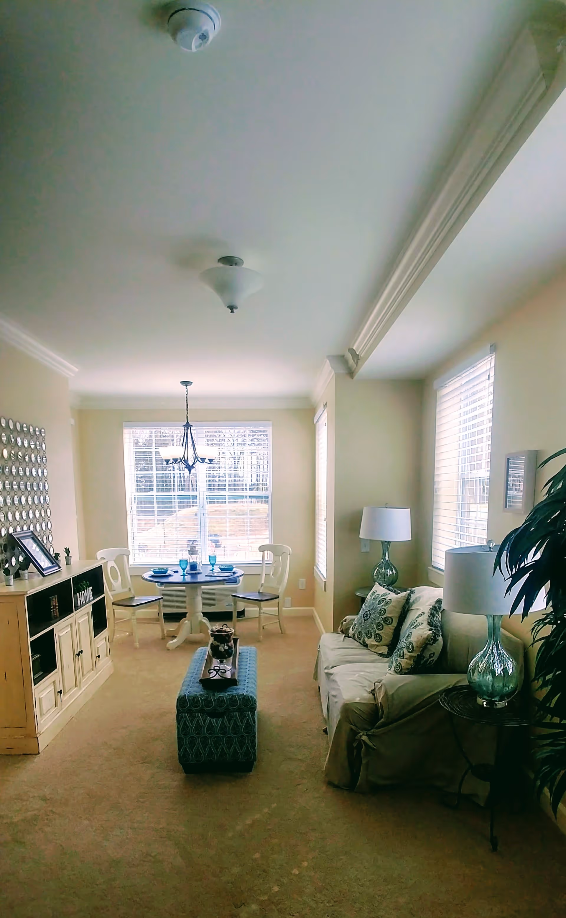 A bright living room and dining area with beige walls and carpet. The living room has a beige sofa with patterned cushions, two glass table lamps on side tables, and a large green plant. The dining area features a round table set with blue glassware and two white chairs, positioned in front of a large window with white blinds. A wooden cabinet with decorative items is on the left side of the room.