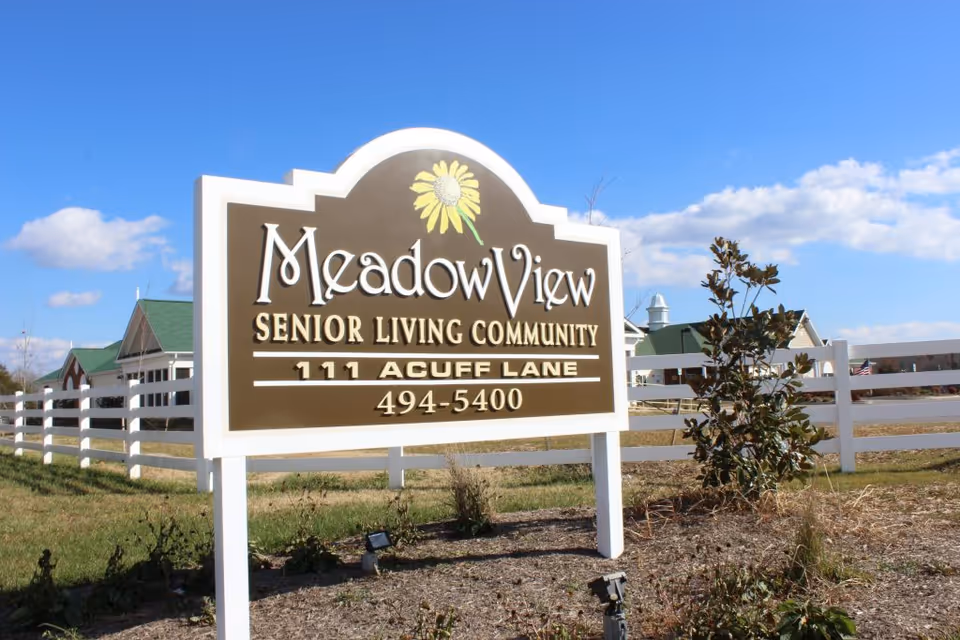 Entrance sign reading 'MeadowView Senior Living Community' with address and phone in front of a white fence and facility buildings under a blue sky.
