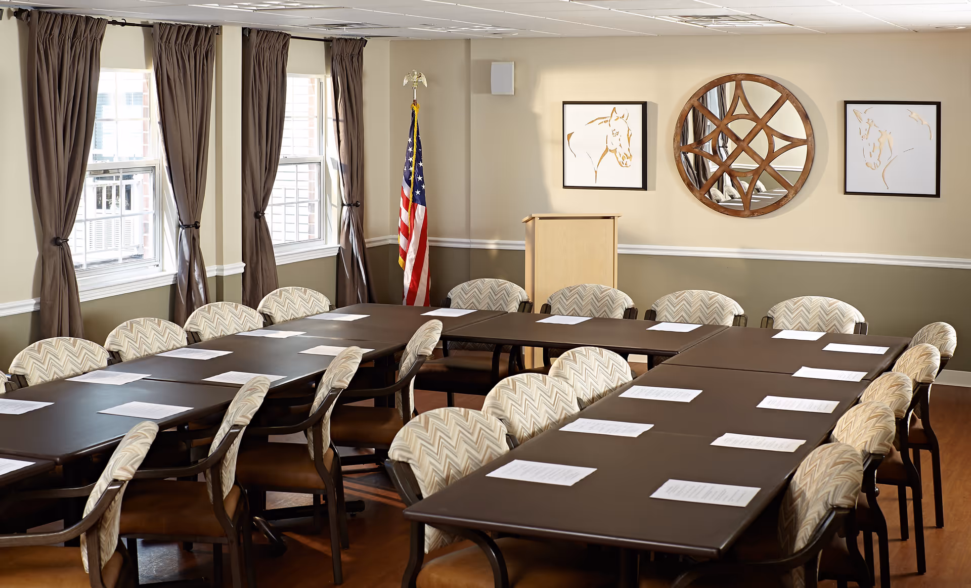 A conference or meeting room with multiple tables arranged in a U-shape, surrounded by cushioned chairs with patterned upholstery. Papers are placed on the tables in front of each chair. The room has three windows with brown curtains, an American flag in the corner, a wooden podium, and three framed artworks on the wall, including a round decorative mirror.