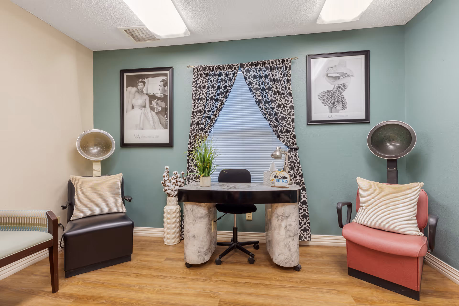 Small salon-like interior with two hooded hair dryers, a central desk and chair, patterned curtains, framed art, and cushioned armchairs.