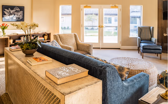 A cozy living room area with a blue sofa, patterned armchairs, a round woven coffee table, and a console table with books and a potted orchid. Large glass doors and windows let in natural light, showing a view of a courtyard outside.