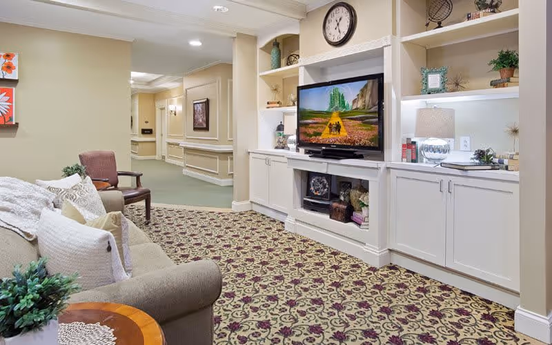 A cozy living room area in a senior living facility featuring a patterned carpet, a beige sofa with multiple pillows, a wooden side table with a plant, a brown armchair, and built-in white cabinetry with shelves. A flat-screen TV is mounted in the cabinetry, displaying a scene from The Wizard of Oz. The walls are painted light beige, and there is a clock above the TV. The hallway leading out of the room is visible in the background.
