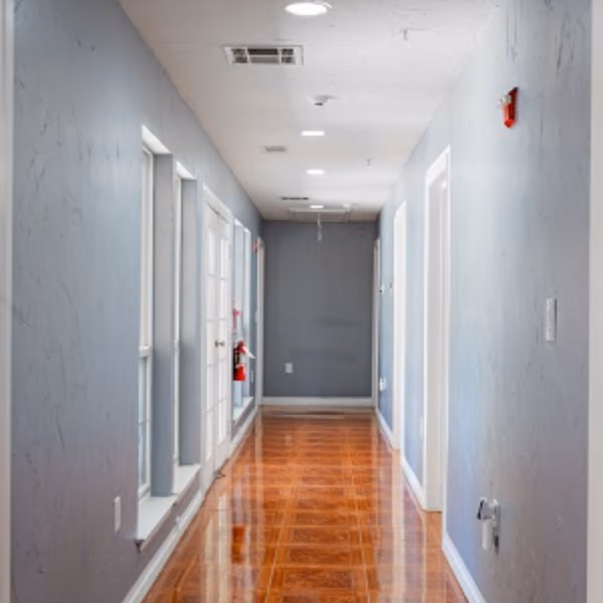 A long, narrow hallway with light gray walls and a polished wooden floor. The hallway has several white doors on the right side and tall windows on the left side. The ceiling has recessed lighting and ventilation vents. A fire extinguisher is mounted on the left wall near the windows.