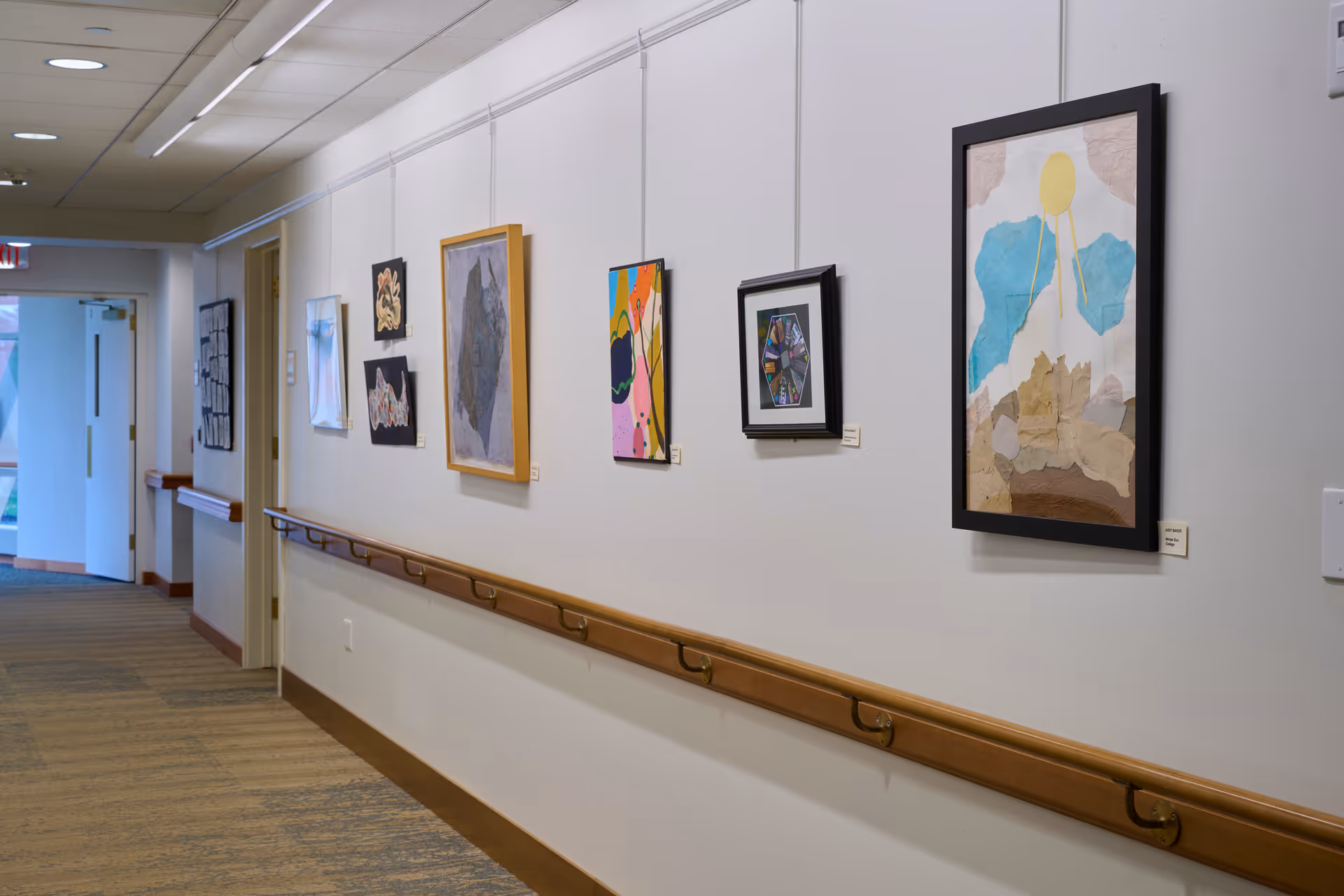 Well-lit interior hallway with framed artwork on a white wall, wooden handrails, and a carpeted floor.