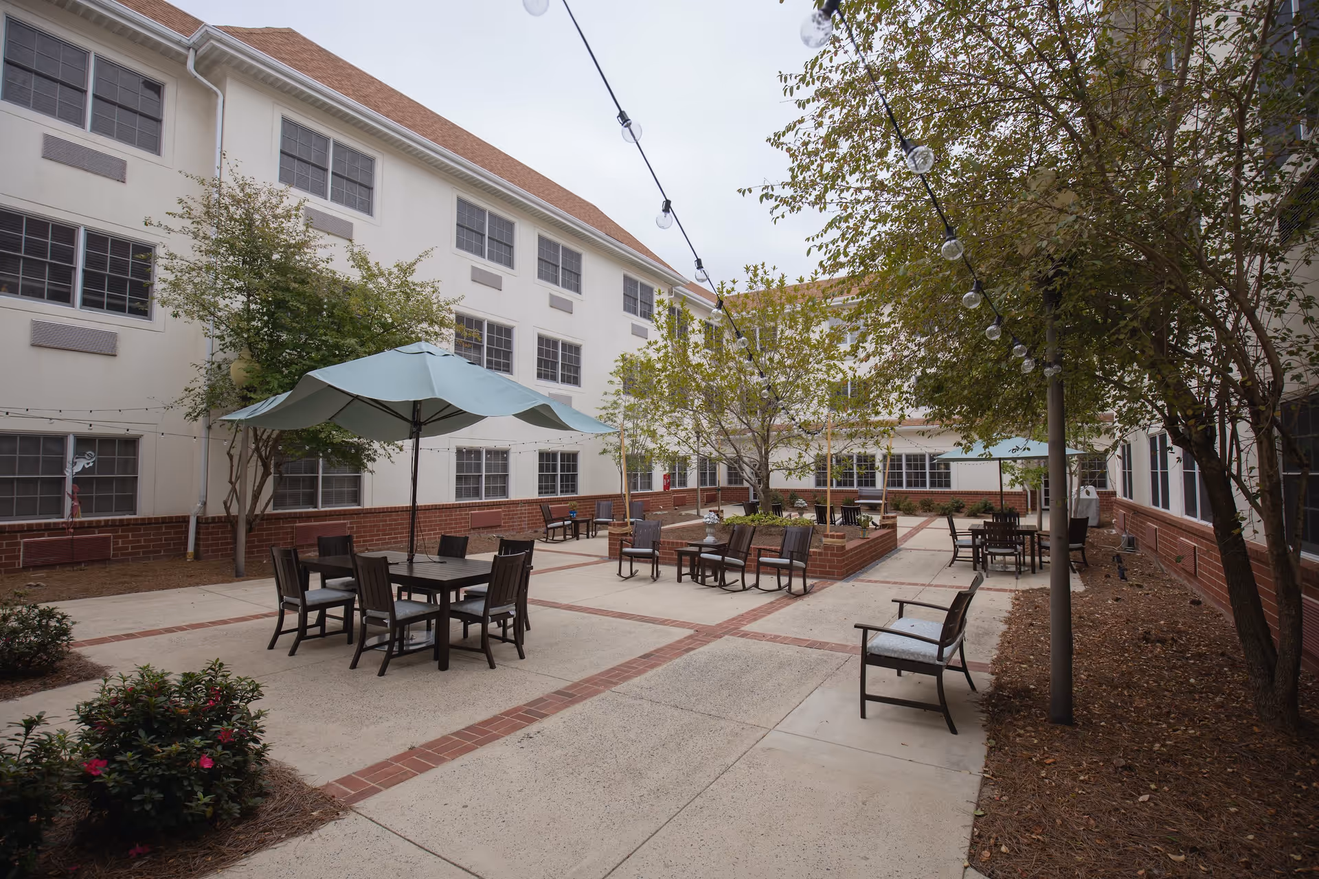 Outdoor courtyard area at The Charlotte featuring multiple seating arrangements with tables and chairs, some shaded by large umbrellas. The courtyard is surrounded by a multi-story building with white walls and red brick accents. Trees and string lights add to the ambiance.