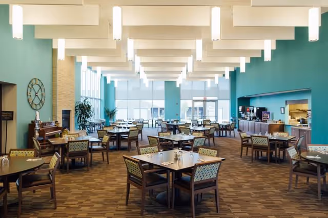 Spacious dining room in a senior living community with multiple tables and chairs arranged neatly. The room features teal walls, large windows allowing natural light, modern ceiling lights, a piano, and a serving area with coffee machines and kitchen access.