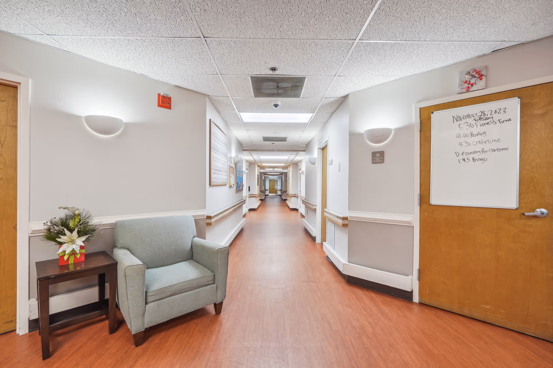 A long hallway in a senior living facility with light-colored walls and wooden doors. There is a light blue armchair next to a small wooden table with a festive floral arrangement. On the right, a door has a whiteboard with a schedule of activities written on it. The floor is a reddish-brown wood-like material, and the ceiling has white tiles with fluorescent lighting.