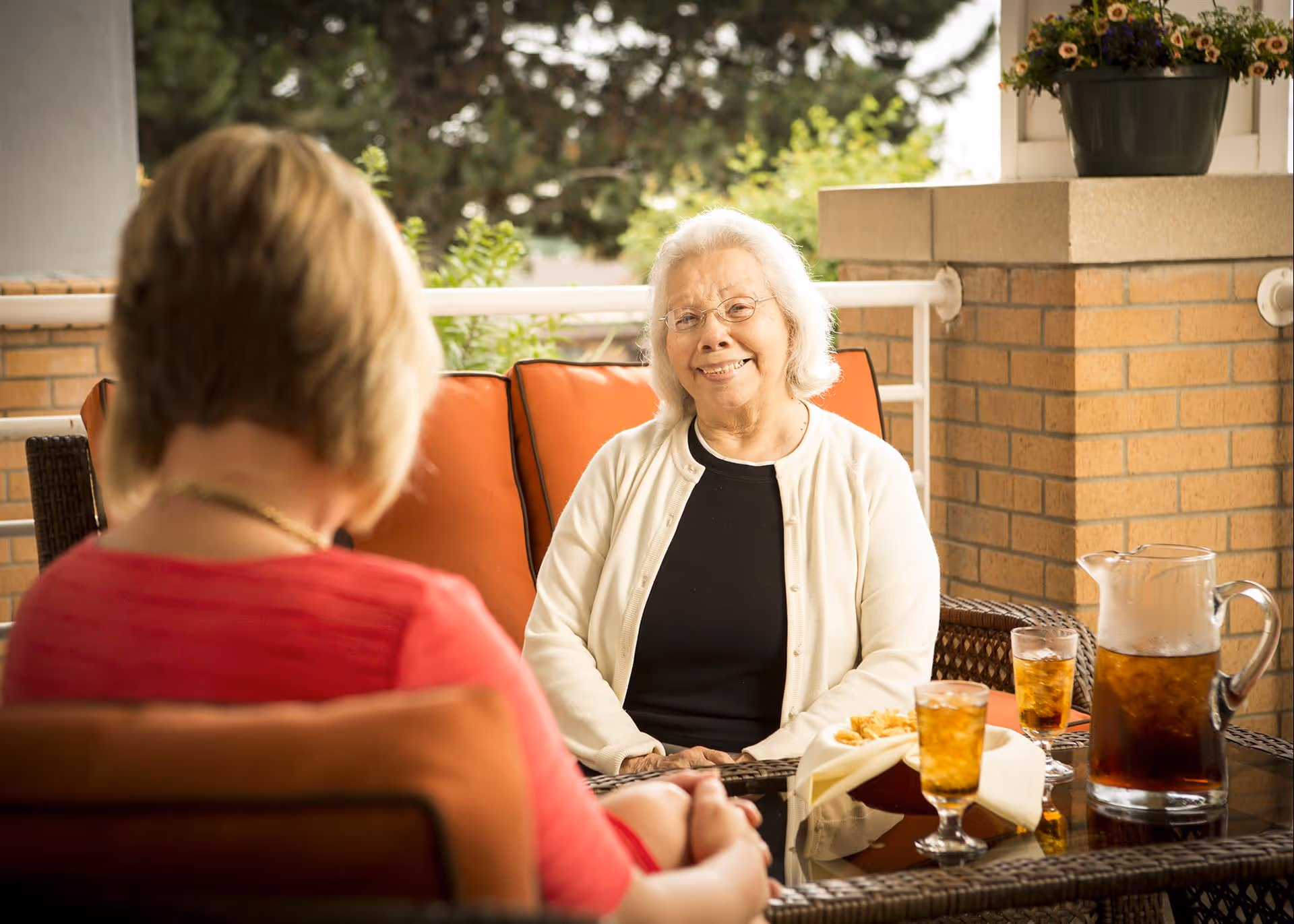 Two elderly women sitting and talking on an outdoor patio with orange cushioned chairs. There is a glass table with a pitcher of iced tea, two glasses of iced tea, and a bowl of snacks. One woman is smiling and wearing glasses, a white cardigan, and a black top. The other woman is seen from behind wearing a red top.