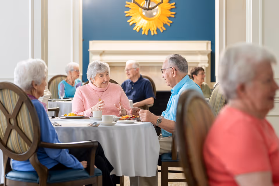 A group of elderly people sitting around a table in a dining room, engaging in conversation and enjoying a meal. The room features a blue accent wall with a decorative sunburst mirror above a fireplace.