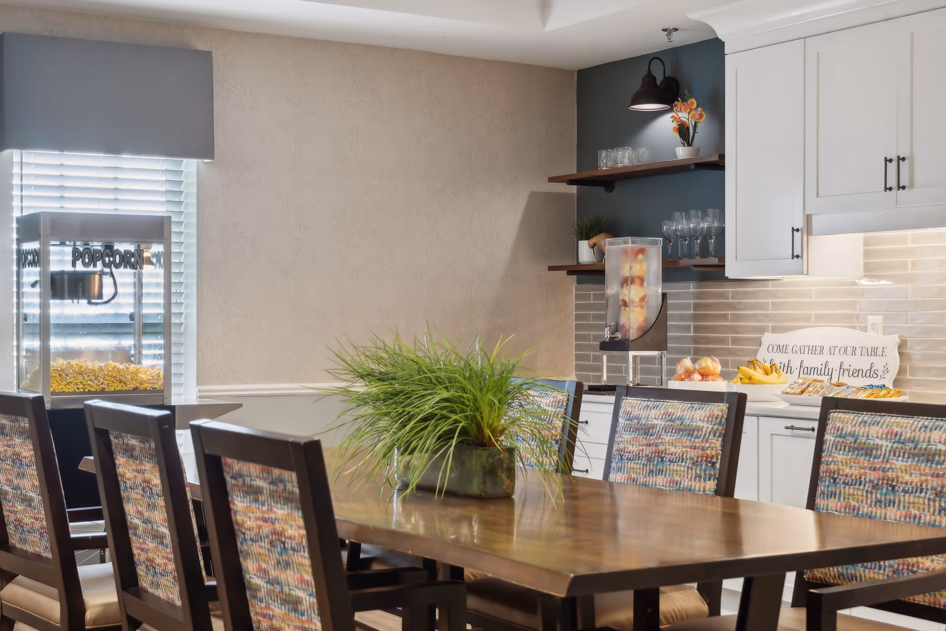Communal dining area with a wooden table and upholstered chairs, a potted plant centerpiece, a popcorn machine by the window and a counter with a beverage dispenser and decorative sign.