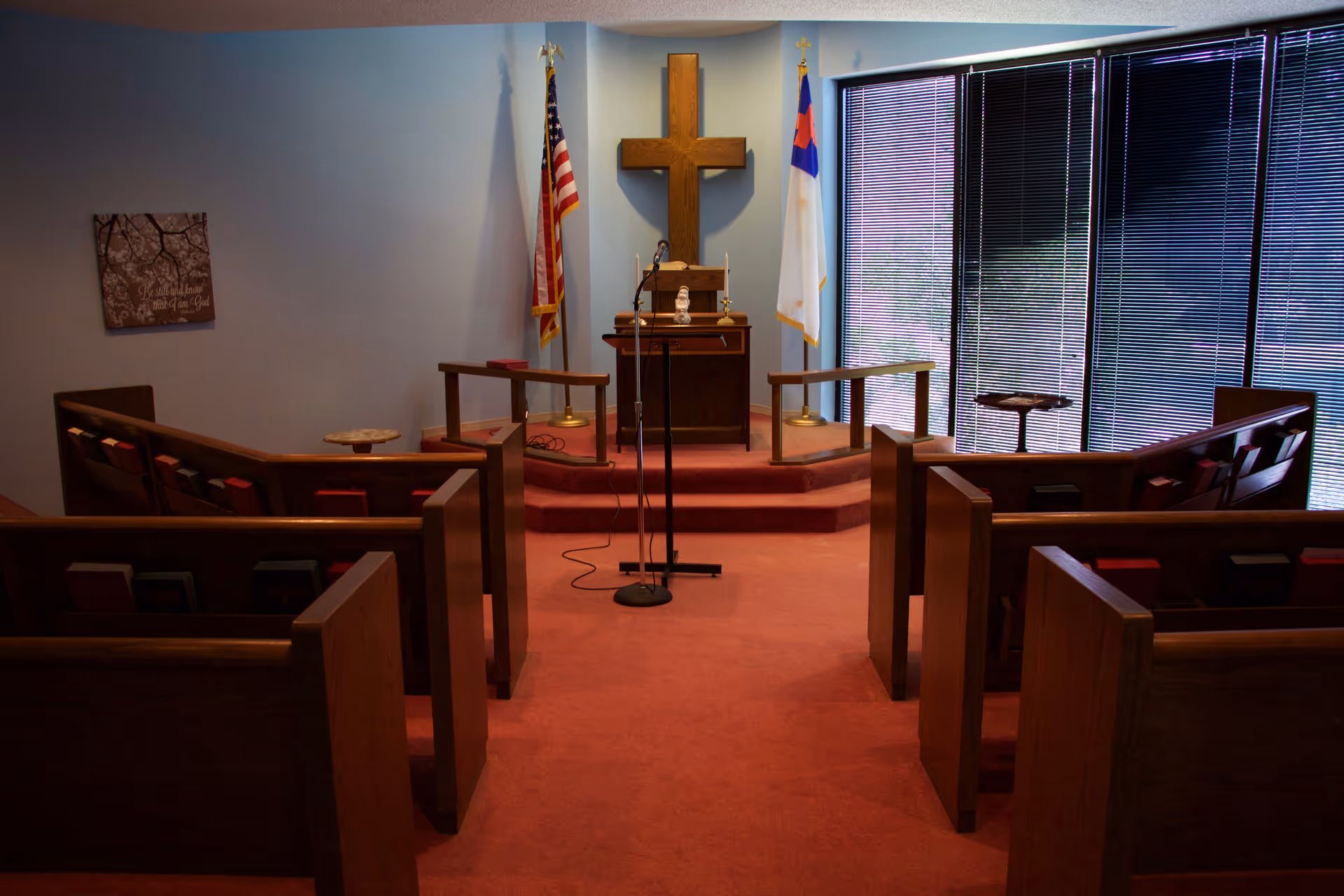 Interior view of a small chapel with wooden pews arranged facing a raised platform with a wooden cross on the wall, an American flag and a Christian flag on either side, a lectern with a microphone, and a small table. The room has red carpet and large windows with closed blinds on the right side.