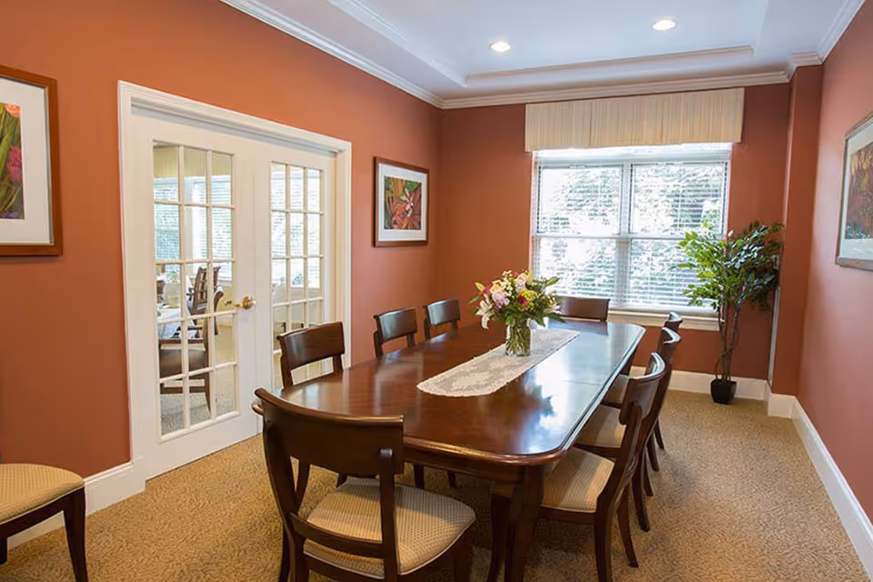 A dining room with a long wooden table surrounded by eight chairs. A vase with flowers sits on a lace runner in the center of the table. The walls are painted a warm reddish-brown color and decorated with framed floral artwork. There is a large window with blinds and a valance, and a potted plant in the corner. French doors with glass panes lead to another room.