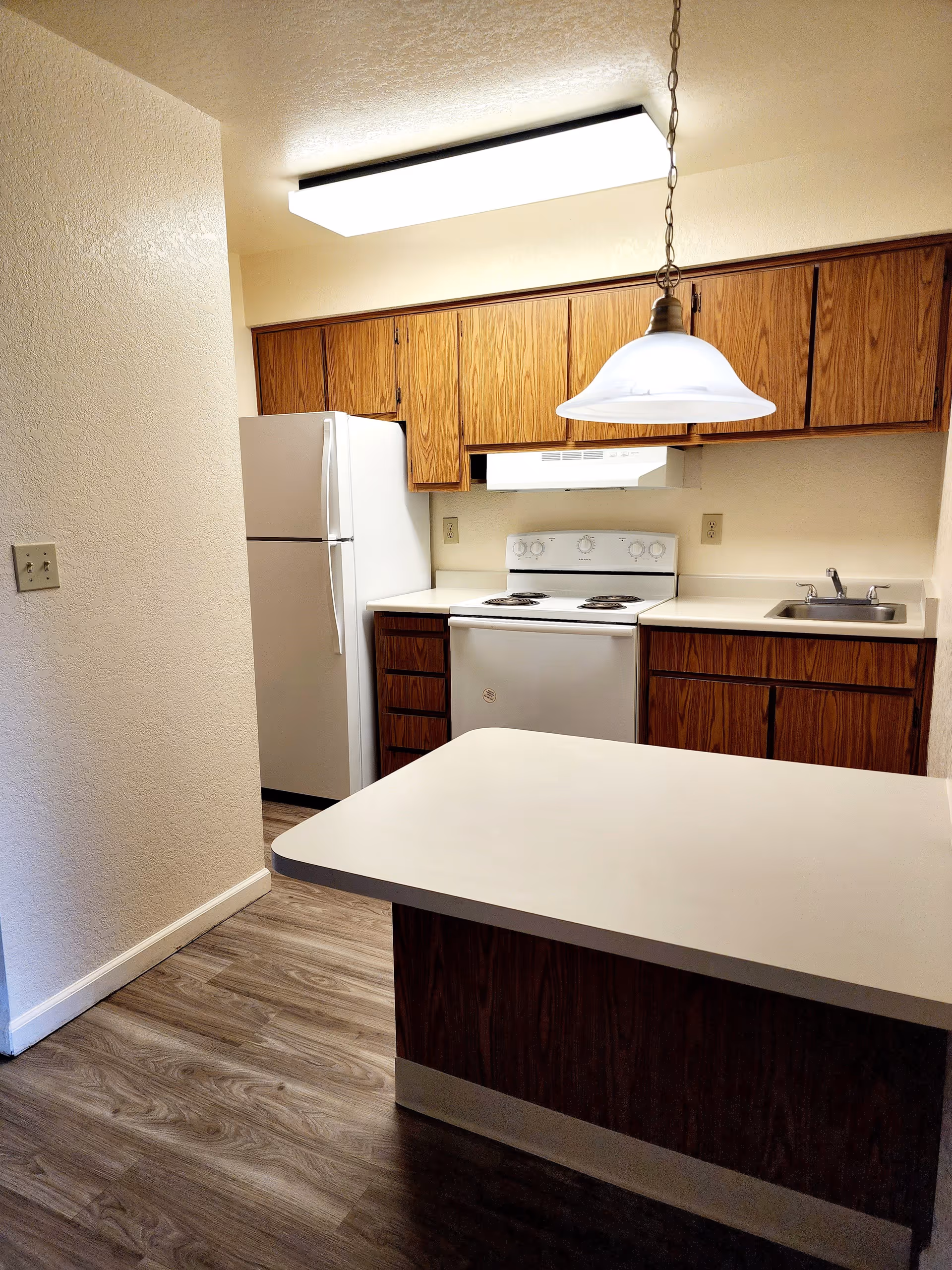 A small kitchen area with wooden cabinets, a white refrigerator, a white electric stove with an overhead range hood, a sink with a faucet, and a countertop island. The floor has a wood-like finish and there is a hanging light fixture above the island.