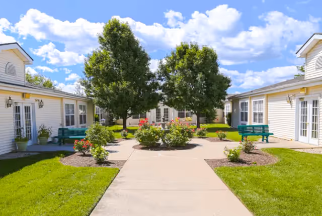 A sunny outdoor courtyard area at a senior living facility with a concrete walkway leading to a central garden bed with flowers and two large trees. The courtyard is surrounded by single-story white buildings with multiple windows and green benches placed on the grass near the buildings. The sky is blue with scattered white clouds.