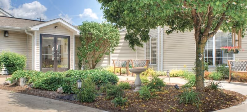 Outdoor courtyard area at Oakwood Terrace featuring a garden with various plants and shrubs, a tree providing shade, a birdbath, benches for seating, and a building entrance with glass double doors in the background under a partly cloudy sky.