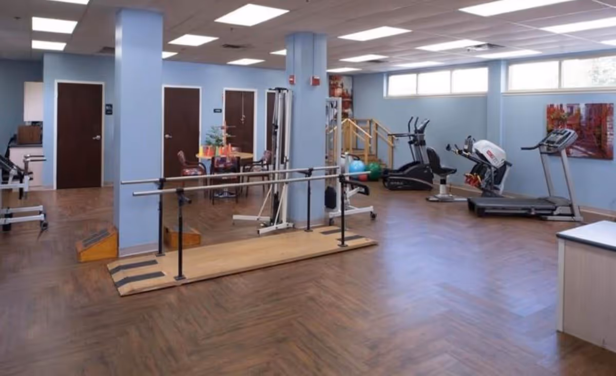 A spacious rehabilitation or physical therapy room with light blue walls and wood-patterned flooring. The room contains parallel bars for walking exercises, various exercise machines, a treadmill, exercise balls, and a small table with chairs. There are several closed wooden doors along one wall and large windows near the ceiling allowing natural light.