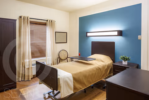A single bed with a beige blanket in a well-lit room featuring a blue accent wall with a modern horizontal light fixture above the bed. Next to the bed is a small nightstand with a potted plant and a movable overbed table. The room also has a wooden dresser, a window with beige curtains and blinds, a round-backed chair, and a framed picture on the wall.