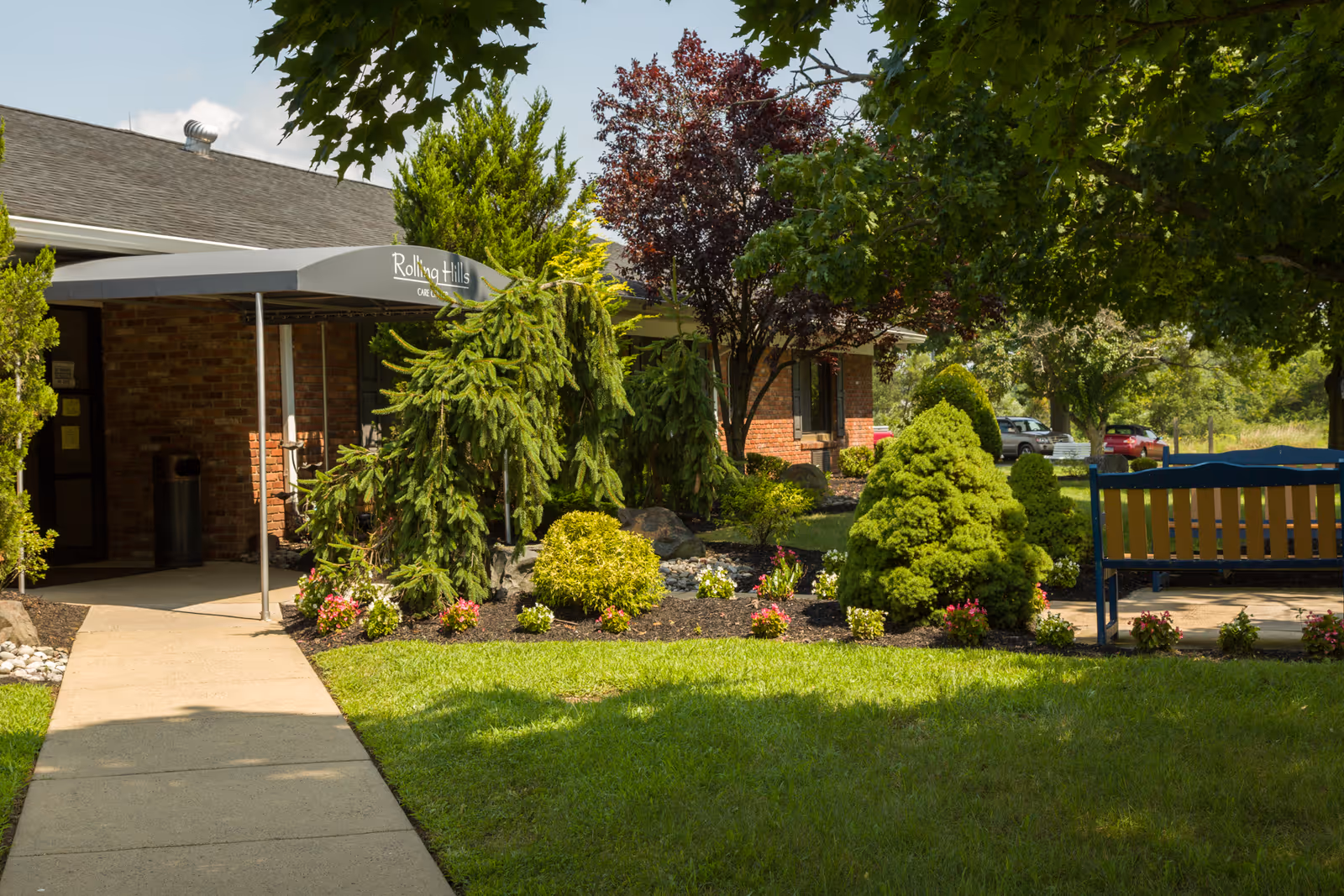 Landscaped front entrance of Rolling Hills Healthcare Center with a walkway, canopy, shrubs and a bench.