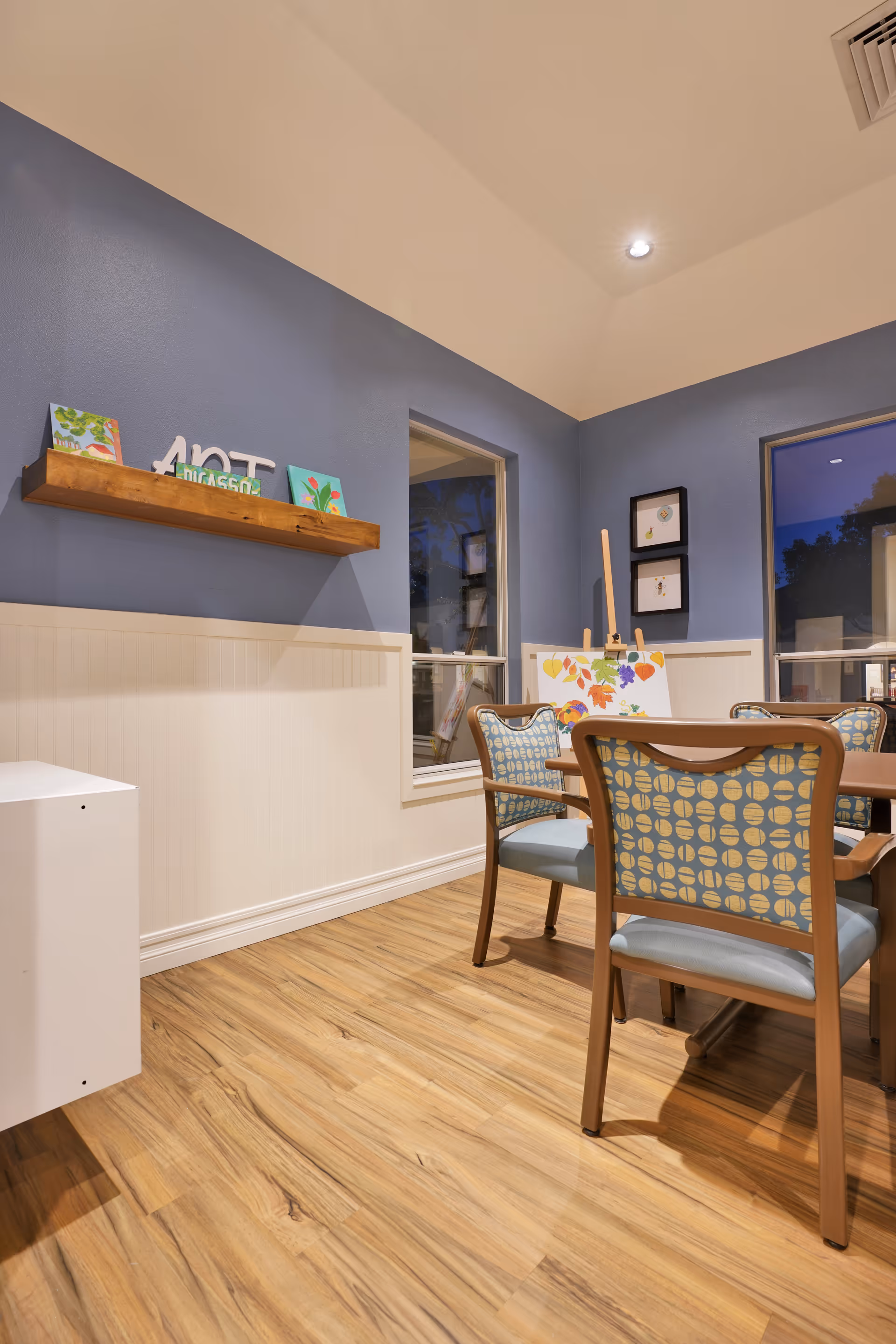 Interior room with blue and white walls, wooden floor, and a wooden table surrounded by chairs with patterned upholstery. A wooden shelf on the wall holds small decorative items and artwork. An easel with a colorful painting is positioned near the window.