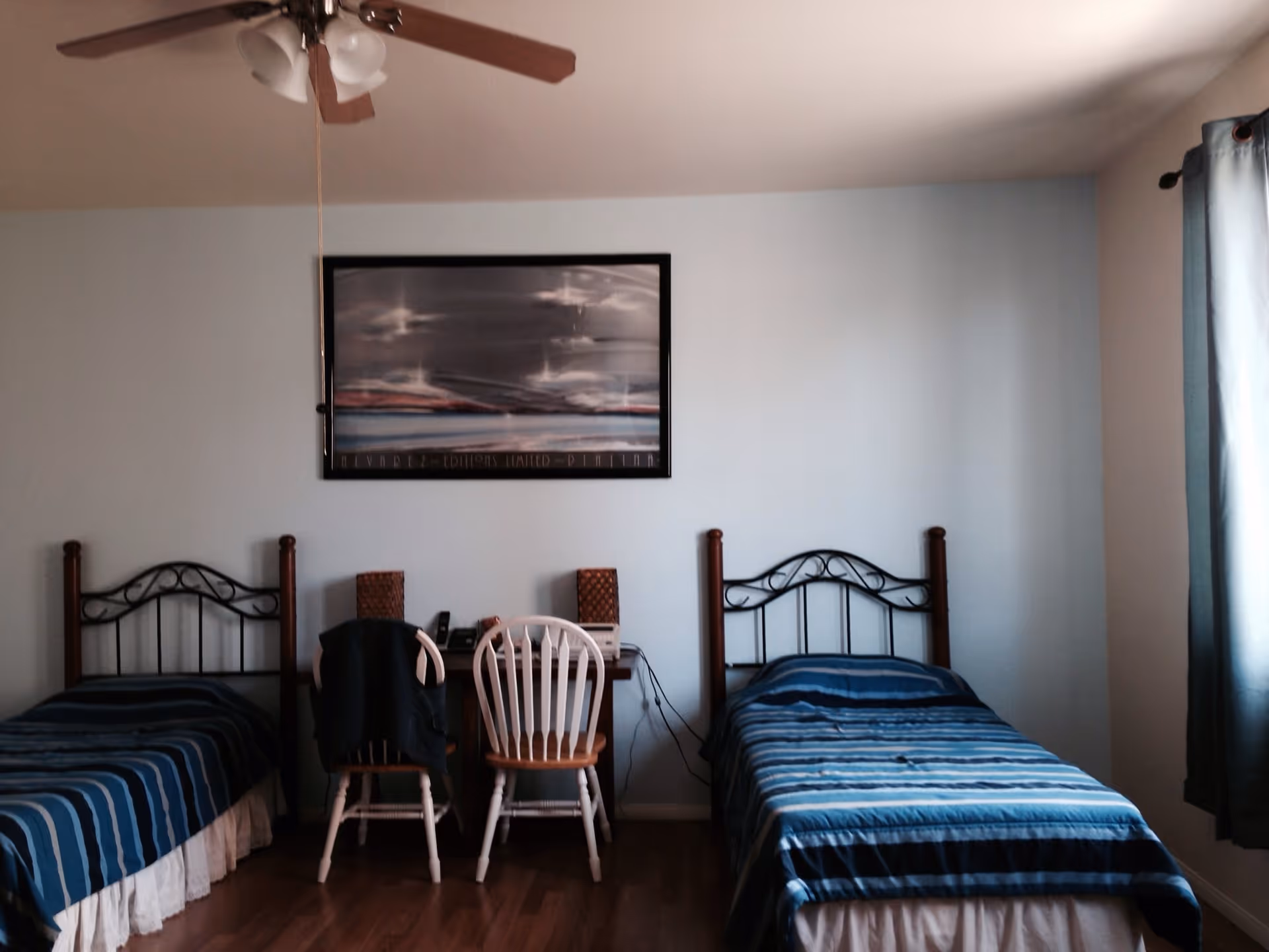 A simple bedroom with two single beds, each with a blue striped bedspread. Between the beds is a small desk with two chairs, one white and one wooden. Above the desk hangs a framed abstract painting. A ceiling fan with lights is visible on the ceiling, and a window with blue curtains is on the right side of the room.