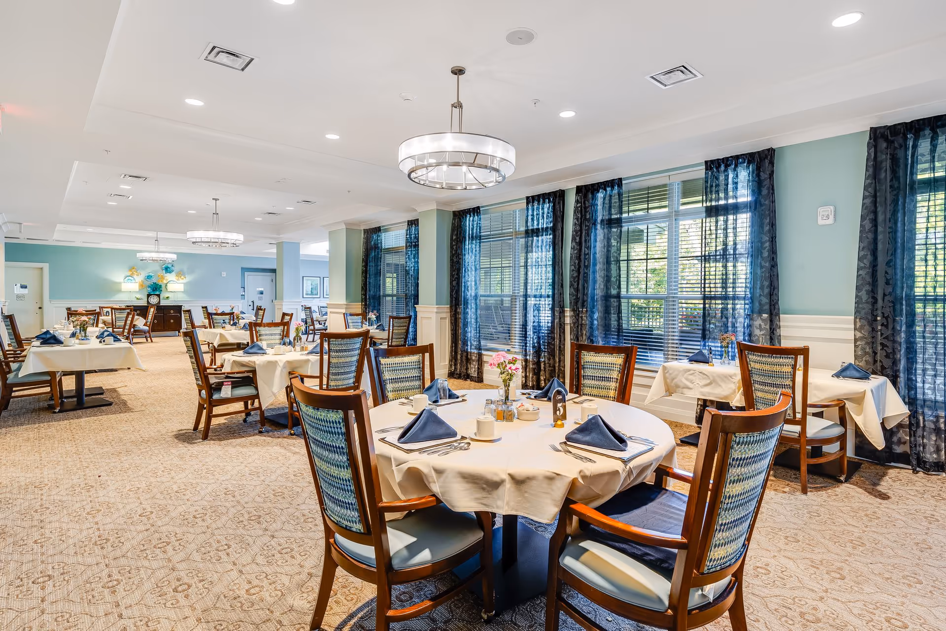 A spacious and well-lit dining room with multiple round and square tables covered with white tablecloths. Each table is set with blue napkins, cups, utensils, and small flower arrangements. The room has large windows with sheer dark curtains allowing natural light to enter. The walls are painted light blue with white wainscoting, and there are modern circular chandeliers hanging from the ceiling.