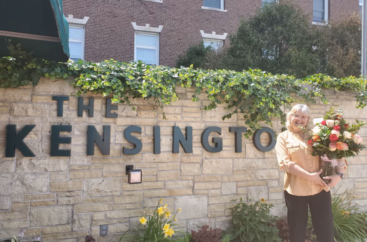 A smiling elderly woman holding a large bouquet of flowers stands in front of a stone wall with green ivy on top. The wall has large black letters spelling out 'THE KENSINGTON'. There are some yellow flowers and green plants at the base of the wall, and a brick building with windows is visible in the background.