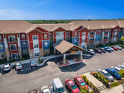 Aerial view of The Welstone at Mission Crossing senior living facility showing a large multi-story building with red and beige exterior walls, a covered entrance, and a parking lot filled with cars in front of the building under a clear blue sky.