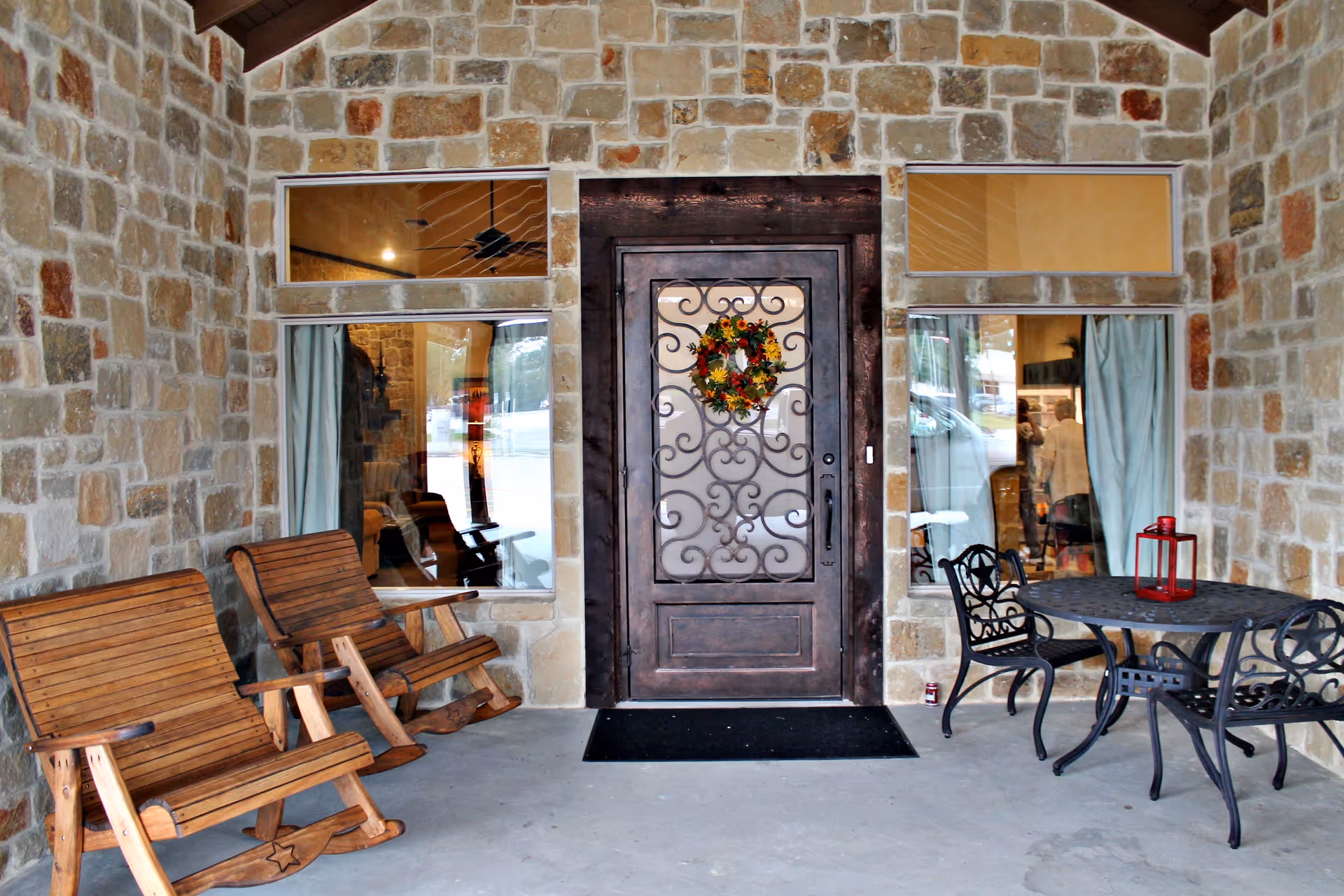 Stone exterior porch area with a decorative dark wooden door featuring a floral wreath. Two wooden rocking chairs are on the left side and a black metal table with two matching chairs and a red lantern on top are on the right side. Two windows with light blue curtains flank the door.