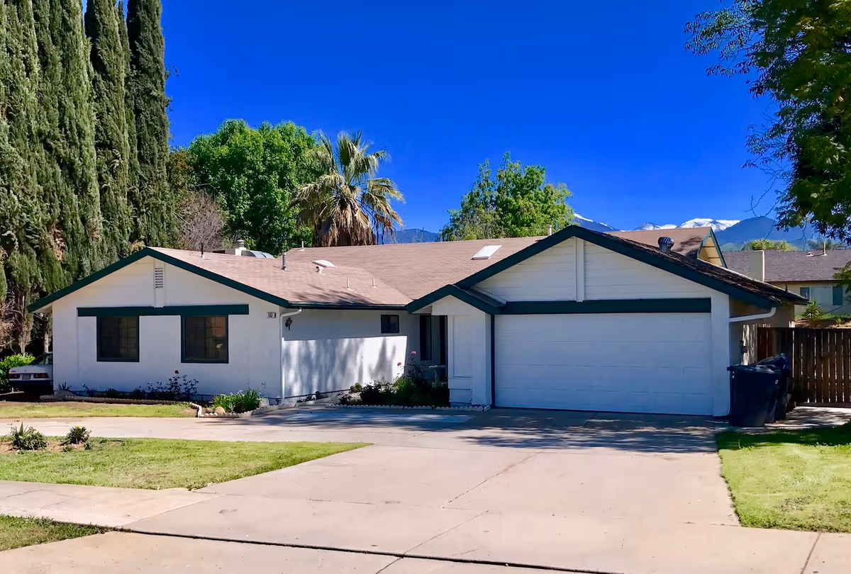Single-story white residential building with an attached two-car garage, driveway, and trees under a clear blue sky.