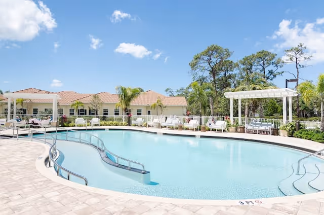 Outdoor swimming pool area with clear blue water, surrounded by a tiled deck with metal handrails. White pergolas and lounge chairs are arranged around the pool. Palm trees and other greenery are visible, with a building featuring a red-tiled roof in the background under a partly cloudy blue sky.