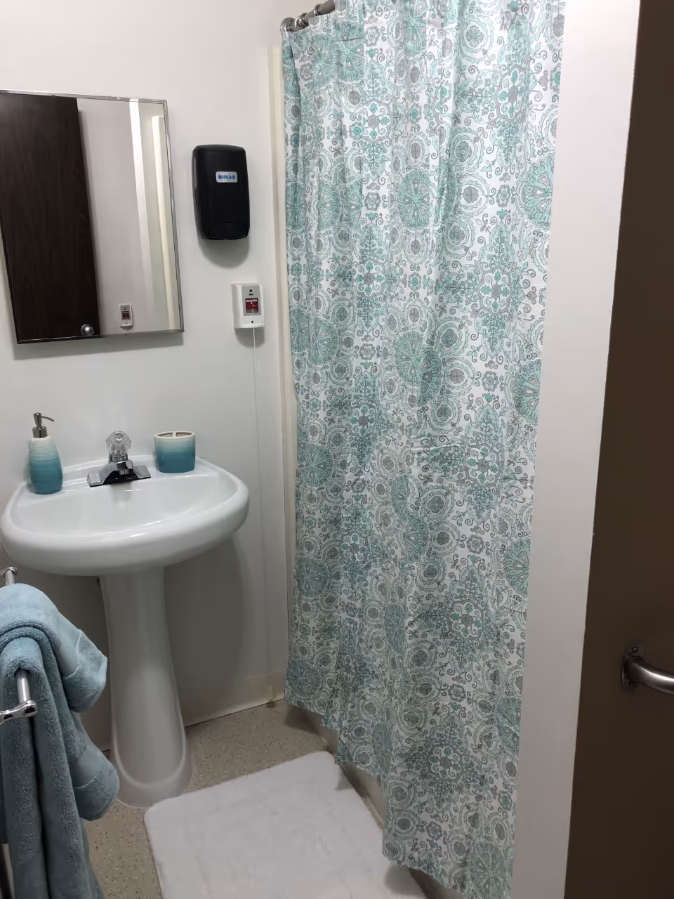 Small bathroom with a pedestal sink, mirror, soap dispenser, teal patterned shower curtain and matching towels.