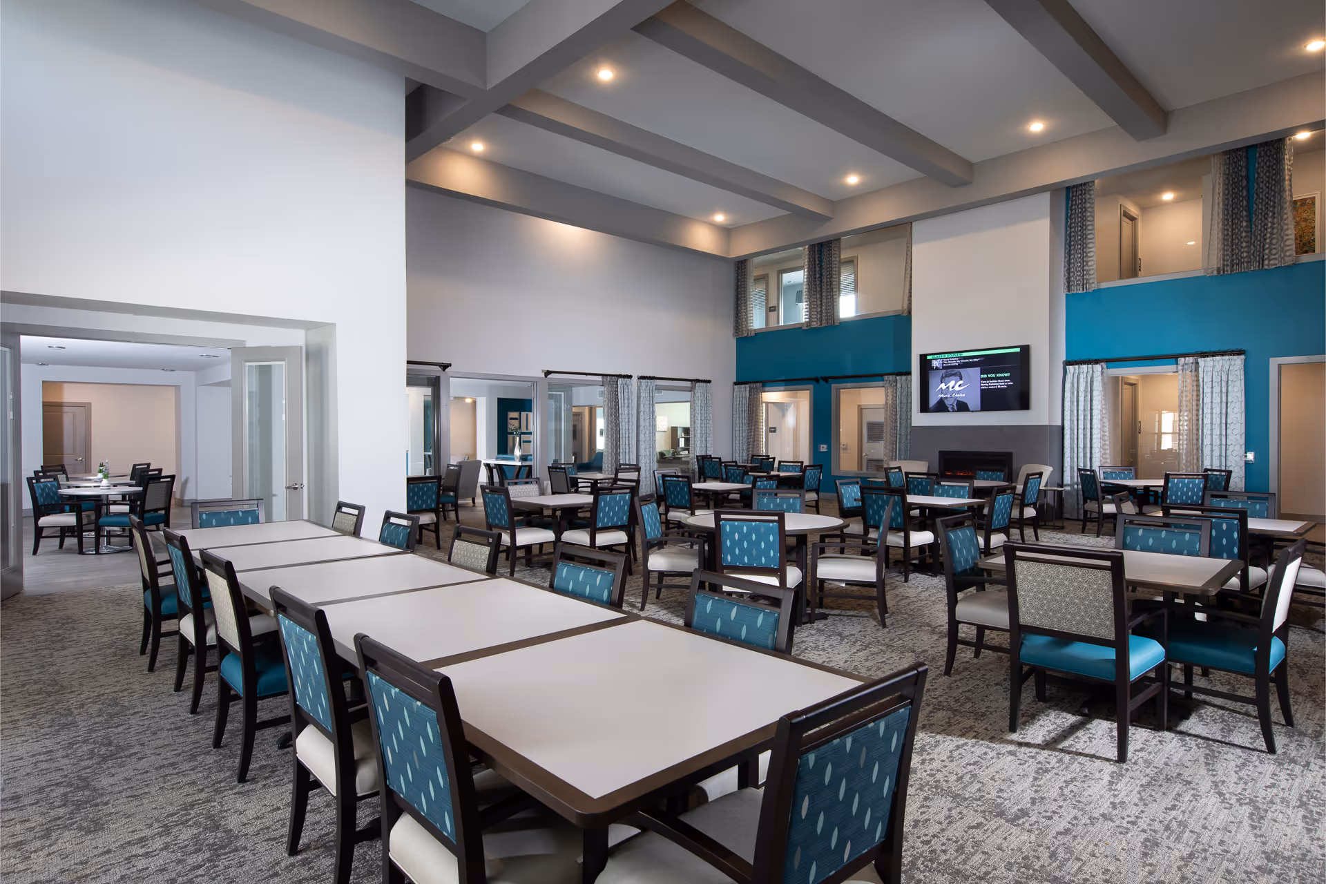 Spacious communal dining room with multiple tables and teal-upholstered chairs under a beamed ceiling and a wall-mounted TV.