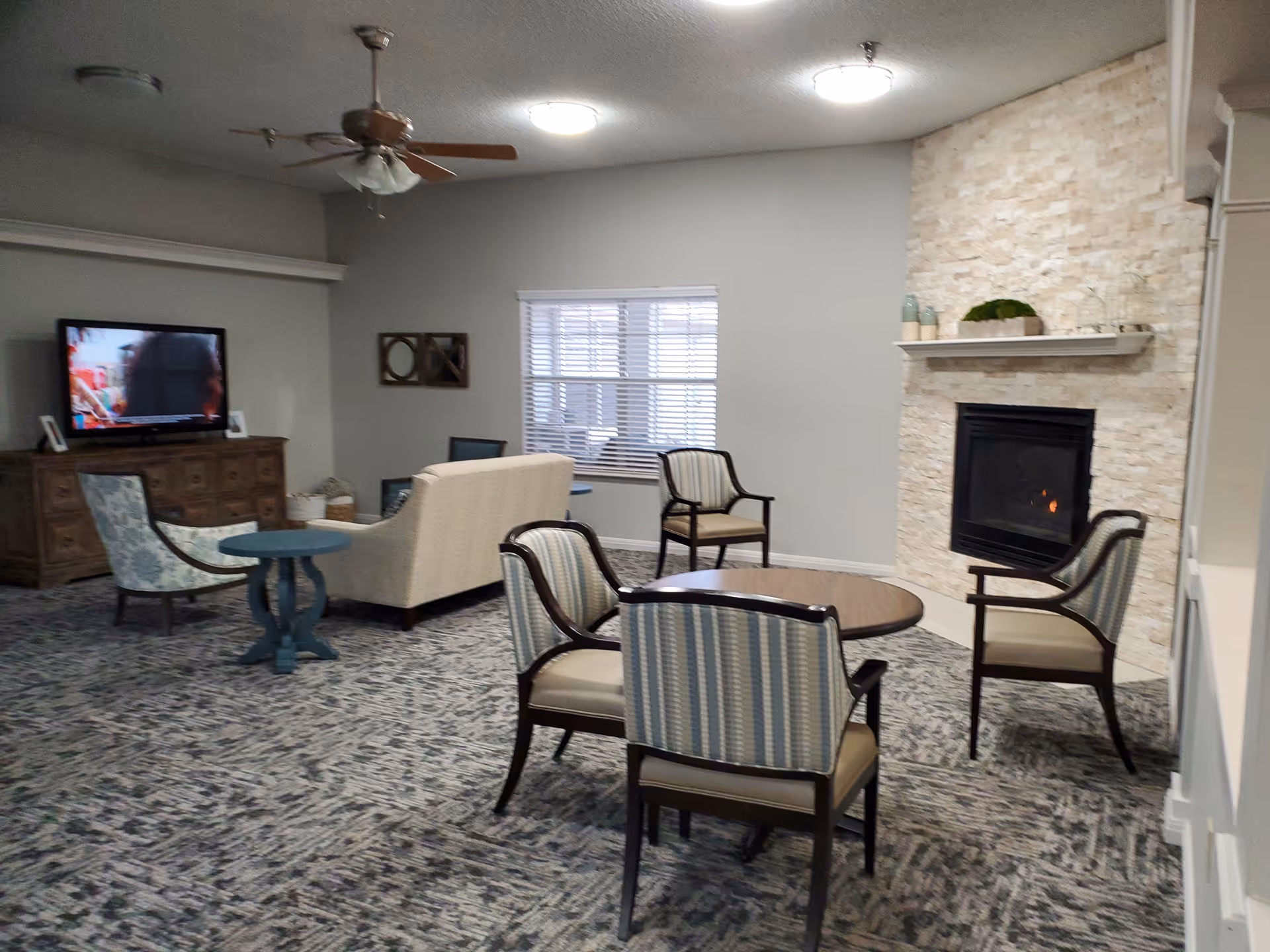 A cozy living room area with a stone fireplace on the right wall, a round wooden table surrounded by four striped upholstered chairs, a beige sofa facing a TV on a wooden cabinet, and a ceiling fan overhead. The room has a patterned carpet and a window with blinds on the back wall.