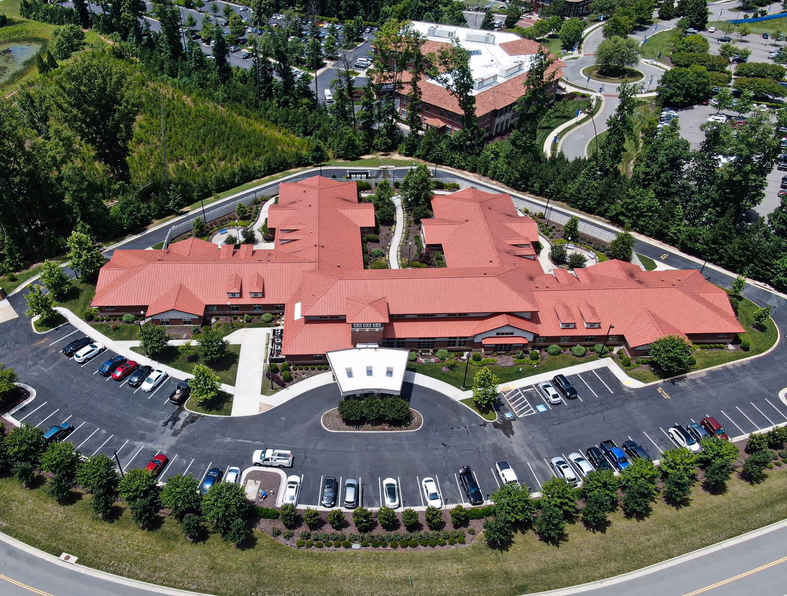 Aerial view of Arden Courts Richmond, a senior living facility with a large building featuring a red roof surrounded by parking lots, trees, and landscaped greenery.