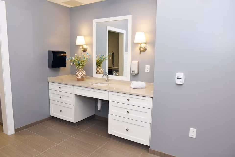 A clean bathroom vanity area with a beige countertop, white cabinets, a rectangular mirror framed in white, two wall-mounted light fixtures with lampshades, a vase with flowers, a rolled white towel, a black paper towel dispenser, and a wall-mounted soap dispenser. The walls are painted light gray and the floor has large beige tiles.