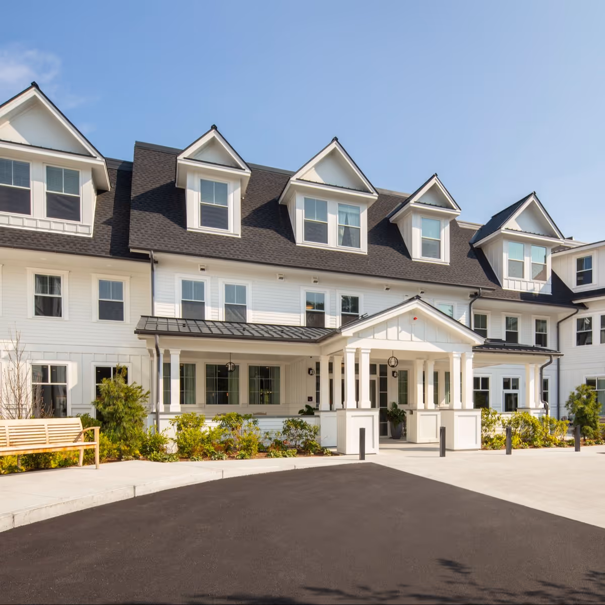 A white three-story senior living building with multiple dormer windows, a covered front entrance, landscaping, and a bench.