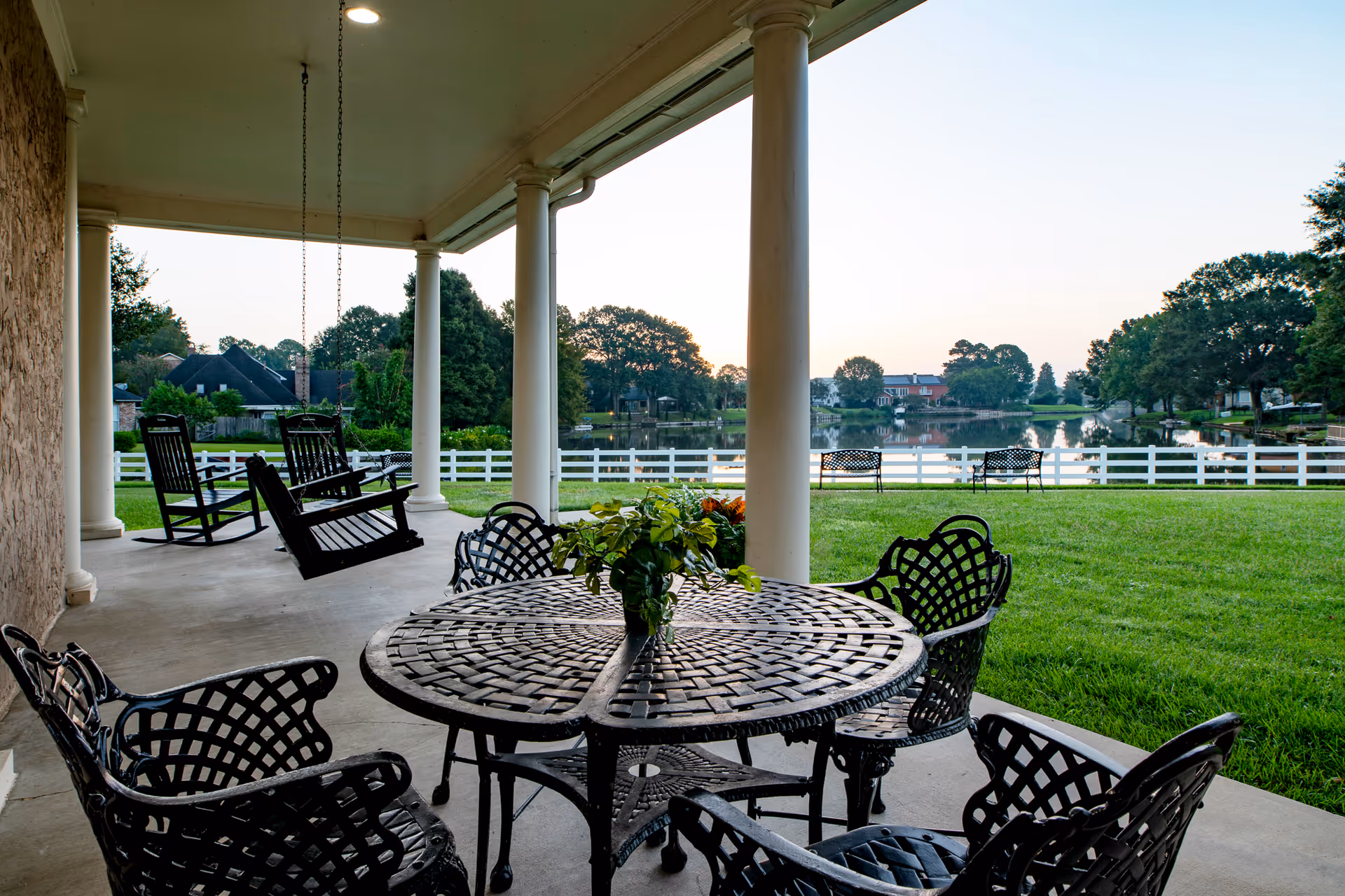 Covered patio area with black metal table and chairs, two black rocking chairs on the left, overlooking a green lawn, white fence, and a calm lake with houses and trees in the background during sunset.