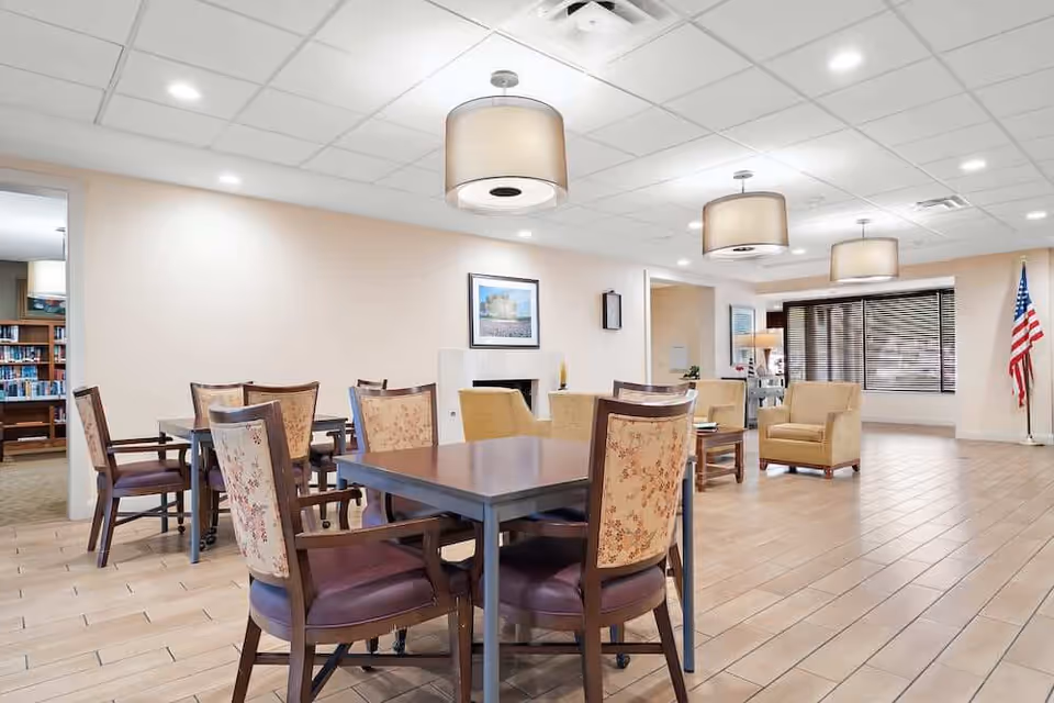 A spacious and well-lit common area in a senior living facility featuring several tables with floral upholstered chairs, beige armchairs, and large hanging ceiling lights. There is a framed picture on the wall, a clock, and an American flag near a large window with blinds. The floor is tiled and the walls are painted light beige.